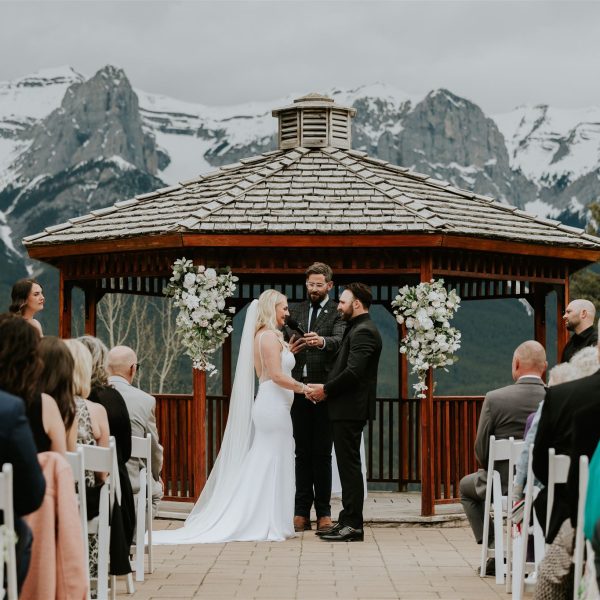 A photographer captures a bride and groom facing each other at an outdoor Canmore wedding ceremony in front of a gazebo, with guests seated and snowy mountains in the background. The officiant stands between them, and floral arrangements decorate the gazebo.
