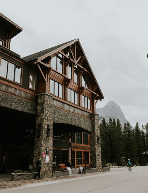 A rustic lodge with stone and wood exterior sits by a road in Canmore, surrounded by tall pine trees. A person stands near the entrance, while another rides a bicycle; a mountain peak rises behind, perfect for an intimate elopement under a cloudy sky.