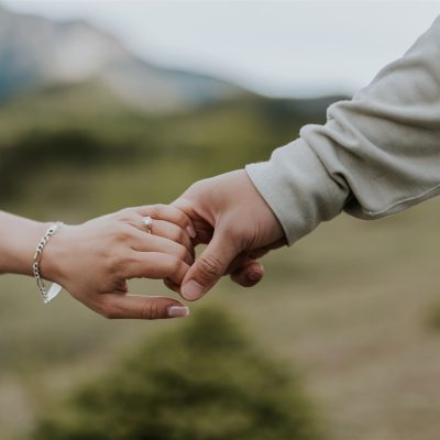 Two people gently holding hands outdoors, with blurred greenery and mountains in the background—a perfect moment for a Calgary wedding photographer to capture. One person wears a gray sleeve, the other a bracelet and ring.