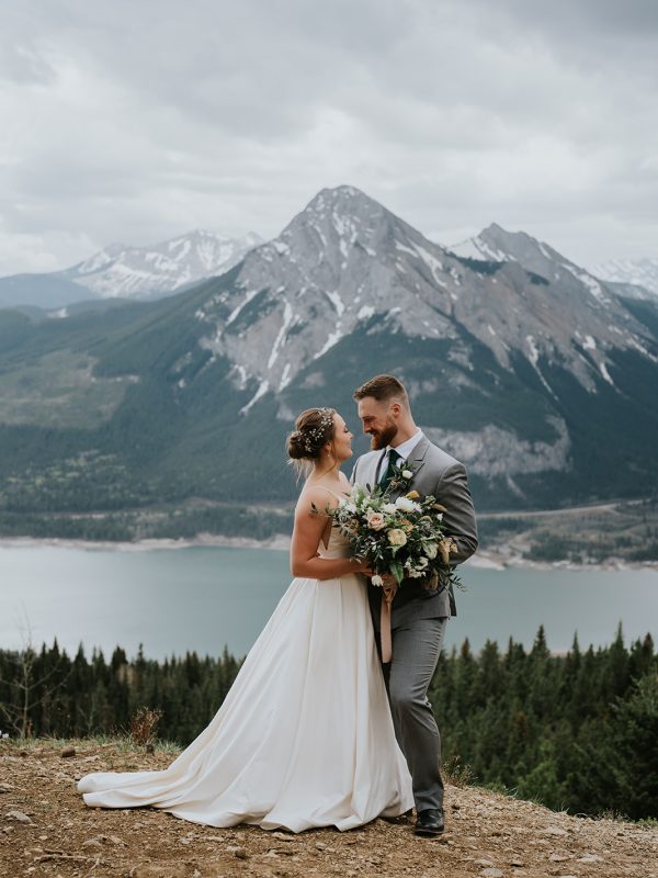 A bride and groom stand together on a mountain overlook near Banff, embracing and smiling at each other. The bride holds a large bouquet, with snow-capped mountains and a lake visible in the background under a cloudy sky.