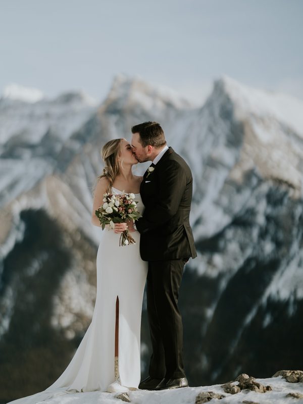 A bride and groom share a kiss outdoors on snowy ground in Canmore, with majestic mountains behind them. The bride holds a bouquet in her white dress, while the groom wears a black suit—perfect for an intimate mountain wedding or elopement.