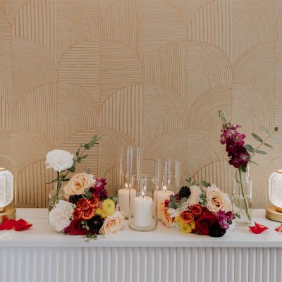 A white table with gold lanterns, elegant floral arrangements in red, white, orange, and pink, scattered rose petals, and glowing candles set against a beige geometric wall—perfect inspiration for a Banff wedding photographer.