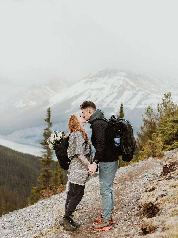 A couple wearing backpacks kisses while holding hands on a mountain trail near Canmore, with a misty snow-capped peak and pine trees behind them—a perfect moment for an elopement photographer to capture. The woman carries a bouquet in her backpack.